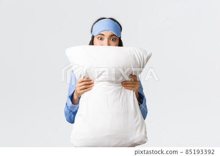 Shocked asian girl in sleeping mask and pajama stare in awe, hiding face behind pillow as watching horror movie at sleepover party, standing white background startled 85193392