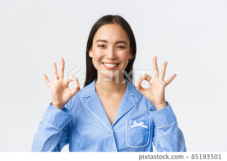 Close-up of satisfied happy asian woman in blue pajama rate nice work, recommend perfect product or service quality, guarantee something as show okay gesture pleased, white background 85193501