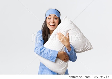 Smiling excited pretty asian girl in blue pajamas and sleeping mask, hugging soft comfortable pillow and looking surprised or amazed at camera at sleepover, white background 85193713