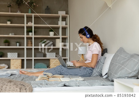 Woman in headphones using laptop, studying online, sitting on bed Woman in headphones using laptop, studying online, sitting on bed 85193842