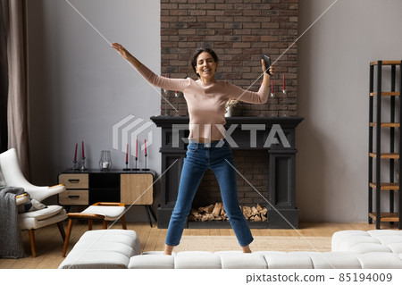 Excited woman holding smartphone jumping on couch at home 85194009