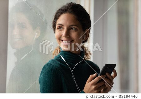 Happy young latin female standing in earphones holding mobile phone 85194446