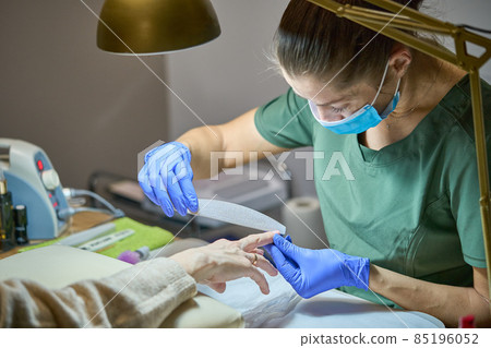 Manicurist filing client nail with a nail file. Manicure process in beauty salon. Master in blue gloves, mask and green uniform under the gold lamp. 85196052