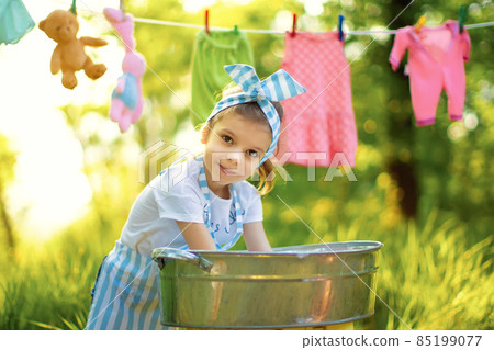 Cute little girl washing her toys in pelvis in checkered apron and headband outdoors 85199077
