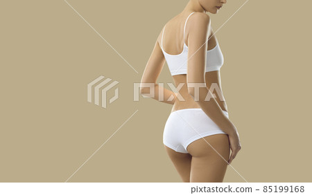 Young model posing in comfy white undies. Woman with beautiful body wearing basic underwear set consisting of soft bra and underpants standing isolated on beige copy space background, view from behind Young model posing in comfy white undies. Woman with beautiful body wearing basic underwear set consisting of soft bra and underpants standing isolated on beige copy space background, view from behind 85199168