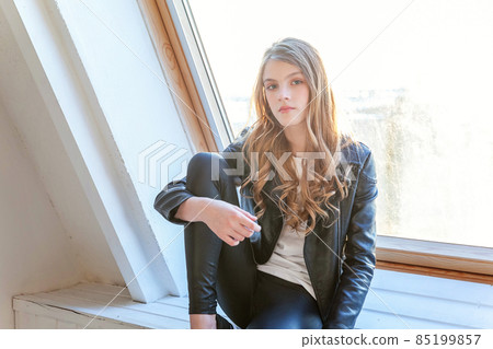 Young teenage girl in leather jacket sitting on modern chair posing in bright light room against white wall background window. Pretty sexy fashion sensual woman dressed in hipster rock style outfit 85199857