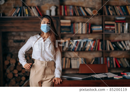 Young woman in medical face mask at home on bookshelves background Young woman in medical face mask at home on bookshelves background 85200119