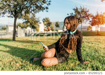 young girl sitting on the grass in a park looking at the smartphone with headphones young girl sitting on the grass in a park looking at the smartphone with headphones 85200121