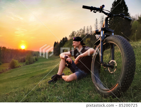 A man is sitting on the grass near a bicycle with thick wheels. Against the background of sunset. 85200725