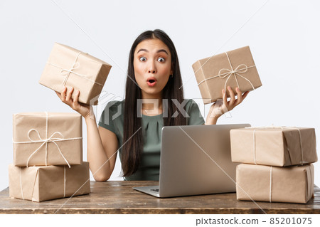 Small business owners, startup and e-commerce concept. Amazed young asian woman open online store, sitting table with laptop, managing clients orders, packing items in boxes for shipping Small business owners, startup and e-commerce concept. Amazed young asian woman open online store, sitting table with laptop, managing clients orders, packing items in boxes for shipping 85201075