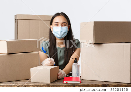 Small business owners, e-commerce and covid-19 preventing virus concept. Smiling asian businesswoman in medical mask and gloves, writing down shipping info on box with order, white background 85201136