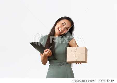 Smiling busy asian shop owner answering phone call, holding mobile on shoulder, prepare package for delivery to client and making notes, checking with customer over phone-call, white background Smiling busy asian shop owner answering phone call, holding mobile on shoulder, prepare package for delivery to client and making notes, checking with customer over phone-call, white background 85201216
