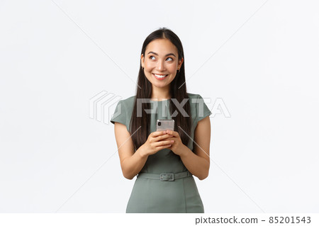 Small business owners, women entrepreneurs concept. Dreamy smiling stylish asian blogger, looking upper left corner thoughtful, creating content, thinking while texting message, using smartphone 85201543