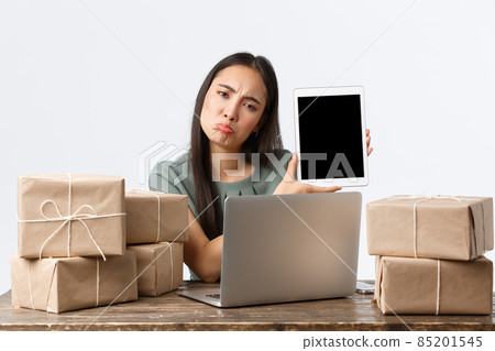 Small business owners, startup and e-commerce concept. Disappointed asian woman pouting upset, showing bad stats on digital tablet screen, using laptop, packing orders in boxes for shipping Small business owners, startup and e-commerce concept. Disappointed asian woman pouting upset, showing bad stats on digital tablet screen, using laptop, packing orders in boxes for shipping 85201545