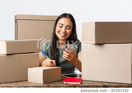 Small business owners, startup and e-commerce concept. Smiling asian businesswoman, online store owner writing shipping addresses on boxes with orders, checking info in mobile phone 85201582