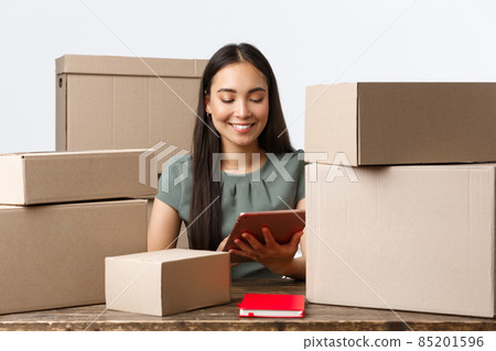 Small business owners, startup and e-commerce concept. Successful asian businesswoman work home, manage internet store from digital tablet, packing orders for shipping, sitting with piles of boxes 85201596