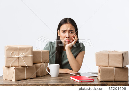 Small business owners, startup and e-commerce concept. Sad and gloomy asian businesswoman pouting as not have clients, waiting for orders, looking distressed sitting near piles of boxes Small business owners, startup and e-commerce concept. Sad and gloomy asian businesswoman pouting as not have clients, waiting for orders, looking distressed sitting near piles of boxes 85201606