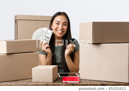 Small business owners, startup and e-commerce concept. Smiling asian businesswoman selling items online, showing credit card and cash, except any payment method, packing orders for shipping 85201612