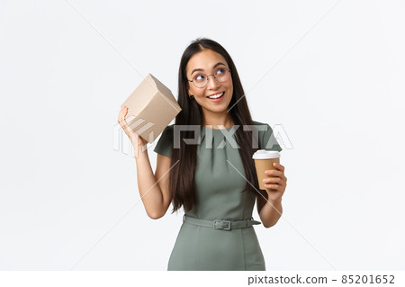 Intrigued smiling asian female drinking coffee, holding mug and shaking box as trying guess what inside, looking curious, receiving parcel from online store, standing white background 85201652