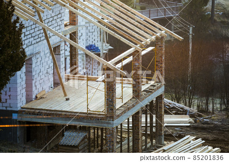 Cottage house building under construction with walls made of hollow foam insulation blocks, wooden roof frame and attached porch terrace with brick columns supporting. 85201836