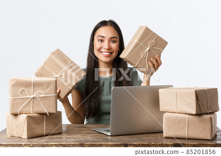 Small business owners, startup and e-commerce concept. Smiling asian woman get lots of orders, sitting table with laptop and packing client purchased goods in boxes for shipping, white background 85201856