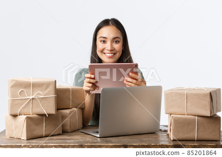 Small business owners, startup and e-commerce concept. Happy asian businesswoman using digital tablet and laptop to contact clients, open internet shop, packing orders in boxes, ready for shipping 85201864