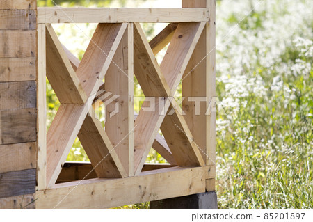 Close-up section geometrical decorative simple detail of new natural not painted wooden planks and bars for balcony, attic fence on blurred blooming summer field background. Construction and design. Close-up section geometrical decorative simple detail of new natural not painted wooden planks and bars for balcony, attic fence on blurred blooming summer field background. Construction and design. 85201897