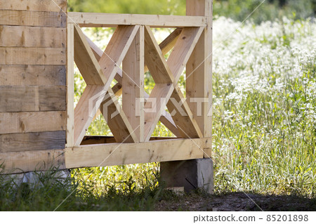 Close-up section geometrical decorative simple detail of new natural not painted wooden planks and bars for balcony, attic fence on blurred blooming summer field background. Construction and design. 85201898