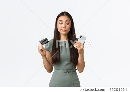 Small business owners, women entrepreneurs concept. Hesitant skeptical asian woman trying new banking application and looking doubtful at smartphone screen while showing credit card Small business owners, women entrepreneurs concept. Hesitant skeptical asian woman trying new banking application and looking doubtful at smartphone screen while showing credit card 85201914