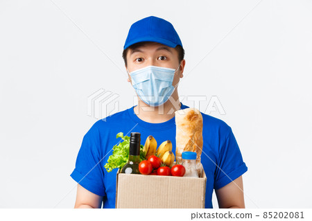 Online shopping, food delivery and covid-19 pandemic concept. Close-up of cheerful asian male courier in medical mask and blue uniform, carry grocery box to client home, standing white background 85202081