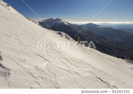Beautiful winter landscape. Steep mountain hill slope with white deep snow, distant woody mountain range panorama stretching to horizon and bright shining sun rays on blue sky copy space background. 85202191