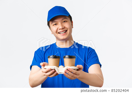 Online orders, takeaway and employees concept. Close-up of cute smiling male courier in blue uniform cap and t-shirt, handing coffee to customer, standing white background 85202404