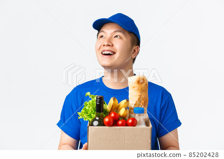 Online shopping, food delivery and shipment concept. Close-up of happy dreamy asian male courier with braces, smiling at upper left corner, carry box with groceries, standing white background Online shopping, food delivery and shipment concept. Close-up of happy dreamy asian male courier with braces, smiling at upper left corner, carry box with groceries, standing white background 85202428