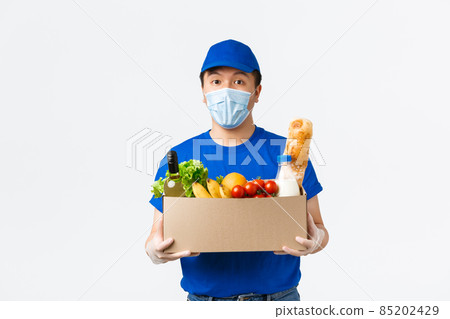 Online shopping, food delivery and covid-19 pandemic concept. Young asian male courier in blue uniform, medical mask and gloves bring grocery order to client home, white background 85202429
