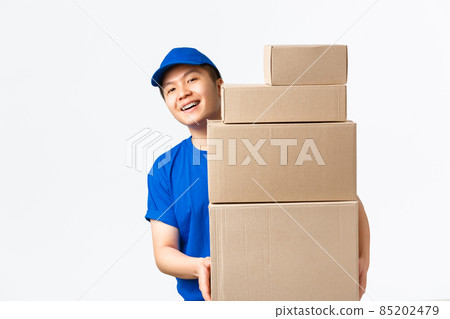Online shopping, fast shipping concept. Smiling happy asian delivery man in blue courier uniform, looking behind boxes with client orders, carry parcels to client home, white background Online shopping, fast shipping concept. Smiling happy asian delivery man in blue courier uniform, looking behind boxes with client orders, carry parcels to client home, white background 85202479