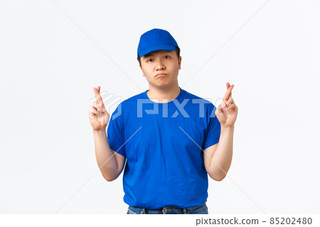 Hopeful gloomy asian delivery man in blue uniform looking at camera, cross fingers good luck, making wish to get raise. Courier praying, want something, anticipating over white background Hopeful gloomy asian delivery man in blue uniform looking at camera, cross fingers good luck, making wish to get raise. Courier praying, want something, anticipating over white background 85202480