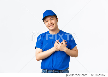 Touched cute asian courier in blue uniform love his clients, care for customer parcels, holding hands on heart and smilig flattered. Delivery guy appreciate praises of his work, white background Touched cute asian courier in blue uniform love his clients, care for customer parcels, holding hands on heart and smilig flattered. Delivery guy appreciate praises of his work, white background 85202487