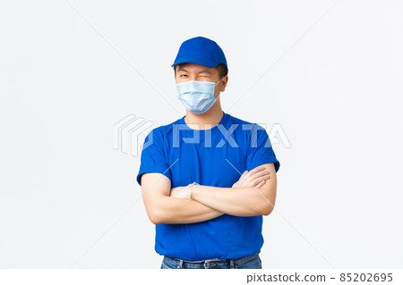 Contactless delivery, covid-19 shipping, preventing virus concept. Sassy friendly asian male courier in medical mask and gloves, blue uniform, winking cheeky and cross arms confident, ensure clients 85202695