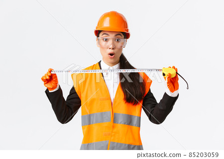 Surprised and excited asian female construction engineer, industrial worker in safety helmet and uniform, measuring layout, holding tape measure and stare amazed at camera, white background 85203059