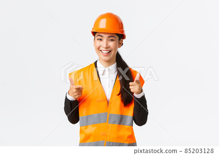 Confident pretty asian female construction manager in safety helmet and reflective jacket, pointing fingers at camera praising nice choice, good work or well done, standing white background 85203128