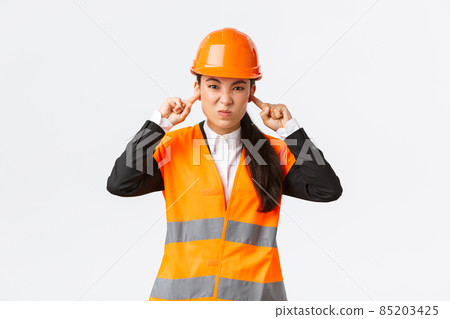 Bothered and displeased asian female chief engineer shut ears and grimacing from awful loud noise at construction area, wearing safety helmet, complaining on bothering sound, white background 85203425