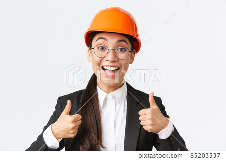 Excited happy female asian engineer, construction manager in safety helmet and business suit showing thumbs-up in rejoice, approve very good work, praise well done job, white background 85203537