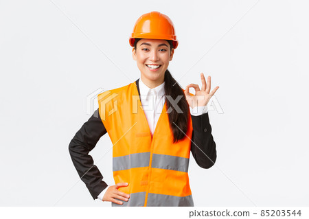 Confident professional asian female architect in safety helmet assure quality and work in time, showing okay gesture and smiling determined, standing assertive, ensure and guarantee something 85203544