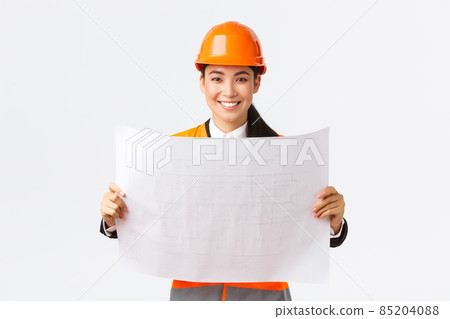 Excited smiling asian female engineer at construction area study blueprints, looking pleased while reading documents, building plan, standing in safety helmet over white background 85204088