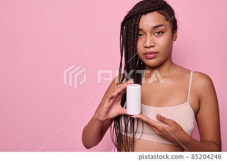 Beautiful African woman with stylish afro dreadlocks in beige underwear holds a jar of moisturizer and and showing it to the camera. Body and skin care concept. Pink background with copy ad space 85204246