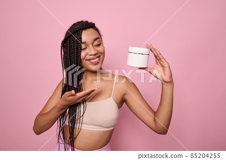 Attractive young African woman in beige underclothes smiles toothy smile pointing on a jar with beauty cosmetic product in her hand, posing against pink background with copy space for ad Attractive young African woman in beige underclothes smiles toothy smile pointing on a jar with beauty cosmetic product in her hand, posing against pink background with copy space for ad 85204255