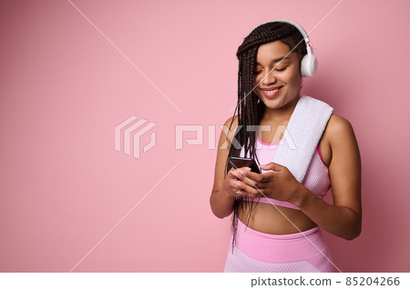 Attractive African American sportswoman in pink tracksuit and wireless headphones, browsing web pages on internet in smartphone, swiping, surfing, isolated over pink colored background 85204266