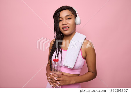 Pleasant young African fitness woman with wireless headphones, dressed in pink tracksuit holding a bottle of water and looking at the camera, isolated on pink background. Sport and recreation concept Pleasant young African fitness woman with wireless headphones, dressed in pink tracksuit holding a bottle of water and looking at the camera, isolated on pink background. Sport and recreation concept 85204267