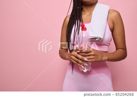 Isolated close-up portrait on pink background of an unrecognizable African woman in pink tracksuit and towel holding a bottle with water, refreshing herself after workout. Copy space 85204268