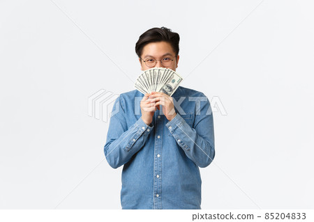 Smiling satisfied asian guy in glasses, hiding face behind money and peeking at camera cunning, earn cash for working freelance, making investment, close deposit, standing white background 85204833